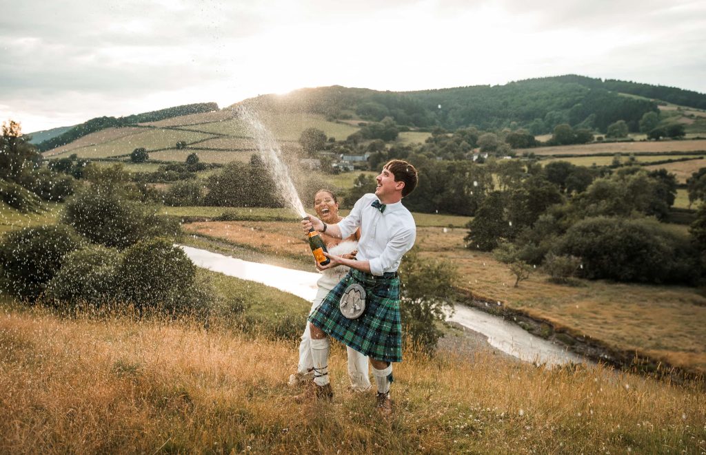 Bride and groom outside near a river. She is in a white dress and he is wearing a white shirt and kilt popping a bottle of champagne
